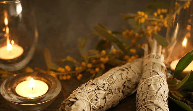 4 Two bundles of sage wrapped in twine next to glowing candles and small green branches with yellow buds on a dark surface.