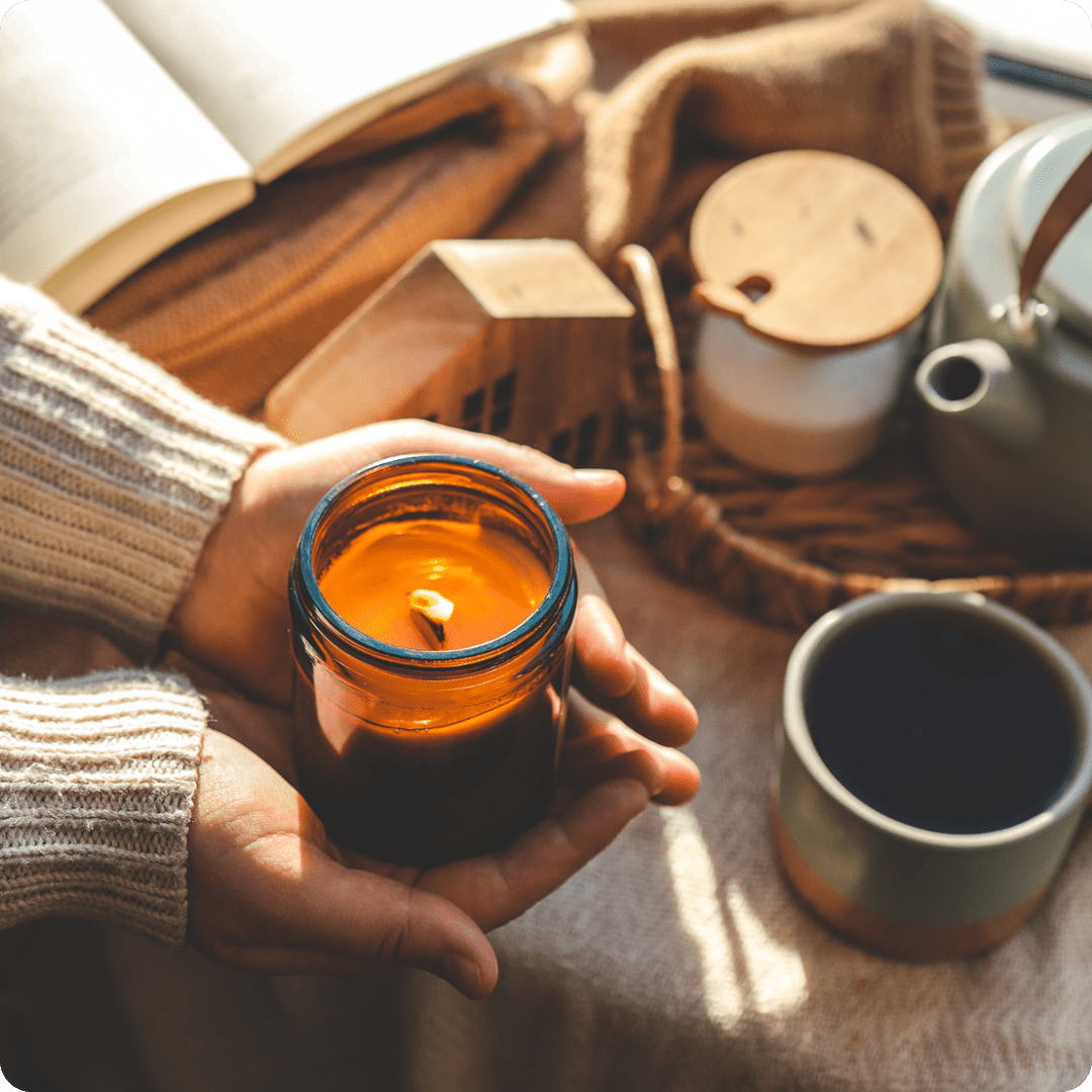 5 Hands holding a lit candle in a glass jar; nearby are a steaming mug, a teapot, and an open book on a cozy blanket.