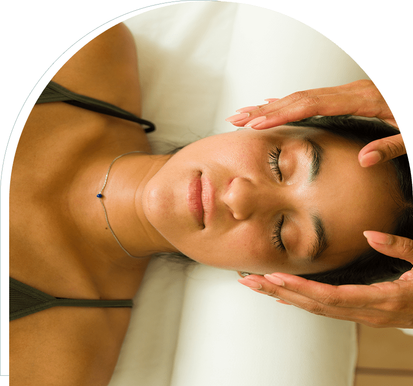 Person receiving a facial massage with eyes closed, lying on a white surface. Two hands gently touch their forehead and jawline.