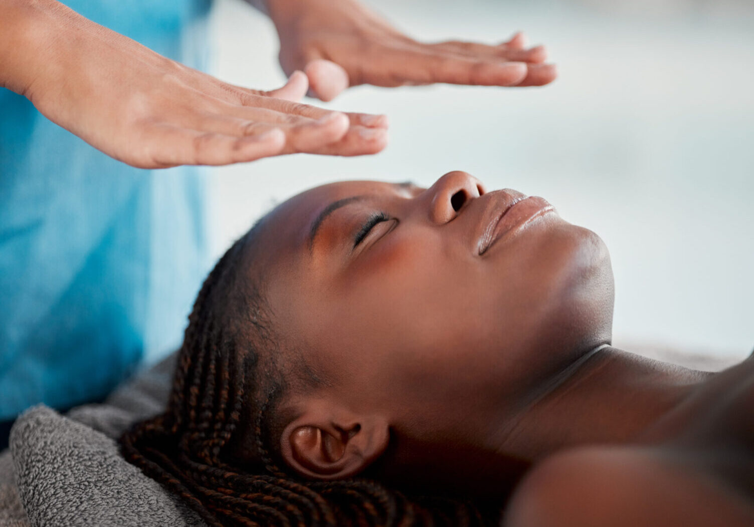 Black woman, reiki massage and luxury spa treatment of a young female ready for salon facial. Skincare, beauty and wellness clinic with client feeling calm and zen from holistic therapy experience Person with closed eyes lies on a massage table while another person holds hands above their face, possibly performing Reiki or energy healing.